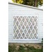 A colorful quilt with a diamond pattern hangs on a white wooden shed, surrounded by greenery.