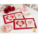 The two completed Tea & Cookies for Two - February placemats, staged on a white table with red roses, white teacups, a pearlescent pink teapot, and frosted heart shaped cookies with pink and red sprinkles.