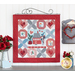 The completed Loveland Cottage wall hanging, staged on a scroll craft holder on a white wall with red flowers and heart related decor.