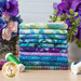 A photograph of a stack of aqua, blue, purple, and multi colored fabrics stacked on a table next to some colorful thread and purple flowers