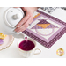 A model pours pink tea from a white teapot into a white teacup. The May placemat serves as the background while the plate of matching cookies peeks into frame from the left.