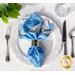 A beautifully arranged blue and white floral napkin is set on an ornate white plate. The napkin is secured with a silver napkin ring. A fork, knife, and a glass of water are placed beside the plate, accompanied by a small green plant in the corner.