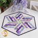 A shot of the completed Bloomerang Table Topper, staged on a white table with coordinating cloth napkins and purple flowers. This photo demonstrates the colors of the fabrics in natural light.