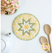 A top down shot of the completed round hot pad in yellow, staged on a white table with coordinating decor like flowers, a white teapot, and wooden spoons.