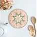 A top down shot of the completed round hot pad in peach, staged on a white table with coordinating decor like flowers, a white teapot, and wooden spoons.