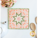 A top down shot of the completed square hot pad in peach, staged on a white table with coordinating decor like flowers, a white teapot, and wooden spoons.