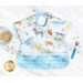 A top down shot of one side of the Toddler bib, showing a white fabric with tossed watercolor portraits of animals and staged with a blue bowl, fork, and cereal.