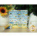 A stack of blue, white, and yellow fabrics with floral summertime motifs, with flowers in a vase and thread and a ceramic beehive