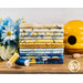 A stack of yellow, blue, and cream fabrics that are in the Bee & Bee Kind FQ set, with flowers and a ceramic beehive.