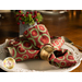 A side shot of a cloth napkin made with red and cream Christmas fabrics in a gold napkin ring in the center of a decorative white plate with gold silverware placed on either side, with a small tin of coordinating holly decor in the background.