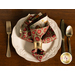 A cloth napkin made with red and cream Christmas fabrics in a gold napkin ring in the center of a decorative white plate with gold silverware placed on either side.