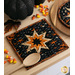 A square quilted pot holder featuring a star design in orange and cream on a black background, with a wooden spoon beside it. There is coordinating seasonal decor in the background as the round and square hot pads sit on a light wooden counter.