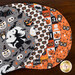 The four completed Oval Placemats colored in gray, orange, and cream fabrics from the Witching Hour collection, slightly fanned out and staged on a brown table.