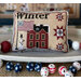 A decorative cross stitch pillow features the word Winter, an American flag, a snowman, and a house design.