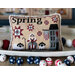 A decorative cross stitch pillow featuring the word Spring, with a farmer, sheep, and a barn in a spring theme.