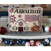 A decorative cross stitch pillow with Autumn text, featuring quilt patterns, a pumpkin, and American flags in a cozy setting.