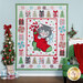 The completed Meowy Christmas quilt, colored in bright green, red, and blue fabrics, hung on a paneled wood paneled wall and staged with coordinating Christmas decor.