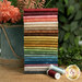 A stack of the fabrics included in the Tea Dye FQ set by Andover Fabrics, with greenery, thread, and flowers in a vase.