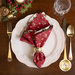 A top down shot of the napkin, staged on a white plate with coordinating cutlery, glassware, and decor, the side with a red fabric with a white snowflake print faced out.