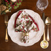 A top down shot of the napkin, staged on a white plate with coordinating cutlery, glassware, and decor, the side with a white fabric with a green holly print faced out.