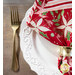 A super close up shot of the red poinsettia fabric of the napkin facing out staged on an ornate white plate next to a gold fork, demonstrating fabric and stitching details.