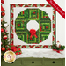 The completed Log Cabin Christmas Wreath in red, green, and white fabrics from the Yuletide Traditions collection, hung on a white paneled wall above a green dresser topped with a blanket of sparkly decorative snow, presents, and a Santa; a decorated Christmas tree peeks into frame from the left side.