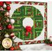 The Wreath at a slight angle, hung on a white paneled wall above a blanket of sparkly decorative snow, presents, and a Santa; a decorated Christmas tree peeks into frame from the left side.