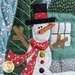 An angled super close up on the center of the wall hanging, featuring a laser-cut waving and dressed up snowman, standing in front of a teal home and tree, demonstrating laser-cut, fabric, stitching, and embellishment details.