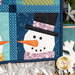 A close up on the lower right snowman, demonstrating fabric, piecing, laser shape, and top stitching details. A decorative tabletop snowflake and silver star can be seen in the corners of the frame.