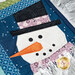 A super close up on one of the snowmen, showing fabric, piecing, laser shape, and top stitching details as a silver garland peeks into frame.