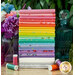 A stack of the fabrics from Tula's True Colors collection for 2025, with a decanter and jars and flowers and thread.