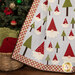 A shot of the bottom left corner of the quilt, demonstrating fabric, border, and binding details. A basket of blankets and a decorated Christmas tree can be seen behind the quilt on the left side of the frame.