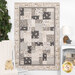 A crib quilt in neutral tones hangs on a wall, surrounded by soft decor with a teddy bear on a chair.