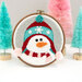 A cheerful snowman in a red and teal hat, framed in an embroidery hoop, amidst pastel trees.