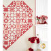 A red and cream patterned quilt hangs on a wall beside a cabinet adorned with flowers.