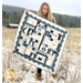 A model holding the completed quilt stands in a snow-covered field, with a forest of trees and small shed in the background.