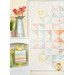 A colorful quilt hangs on a wall near a shelf with stacked fabric and a vase of tulips.