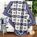 A folded quilt with a geometric pattern in blue and cream draped over a white cabinet next to a floral vase and a potted tree. 