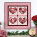 A quilt depicting four heart patterns in pink and red, framed by a floral design, with embellishments.