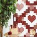 Patchwork quilt featuring heart patterns in red and pink, displayed beside a plant and table with fabrics used in the quilt