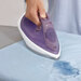 A person uses the push button on a purple iron to spray water on a blue fabric for easy removal of wrinkles.