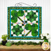 The completed Pieceful Patchwork - March wall hanging, a clover themed project colored in vibrant green, white, and rainbow fabrics hung on a craft wire hanger and staged on a white paneled wall with coordinating greenery and decor.