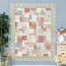 The completed Easy As ABC and 123 Quilt colored in the Just Rosy collection, hung on a blue paneled wall with warm cream and green decor on either side of the quilt.