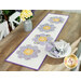 The completed Bloomberg table runner colored in white, yellow, and purple floral fabrics, displayed on a wooden table that's set with a small milk can holding flowers and a white porcelain plate set with coordinating cloth napkins.