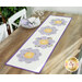 The completed Bloomberg table runner colored in white, yellow, and purple floral fabrics, displayed on a wooden table next to a white porcelain plate set with coordinating cloth napkins.