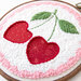 Embroidery of two heart-shaped cherries with green leaves on a circular hoop.