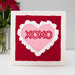 A framed embroidery design featuring a pink and white heart with XOXO on a red background next to roses.