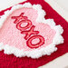 Close up of embroidery design featuring a pink heart with xoxo in red, surrounded by fluffy white edges, set against a red background.