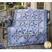The finished Morning Blooms Patchwork Quilt in warm blues, purples, and creams, draped over a wooden bench on a porch surrounded by coordinating décor.  