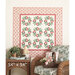 A floral quilt with red and green designs hangs on a wall above a wicker chair with cushions.